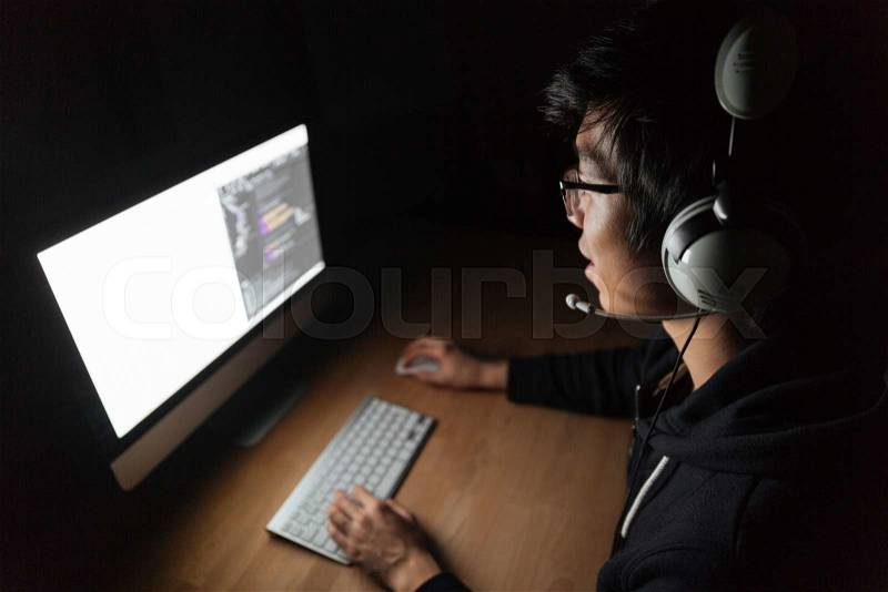 Concentrated asian male gamer sitting and playing computer game in dark room, stock photo