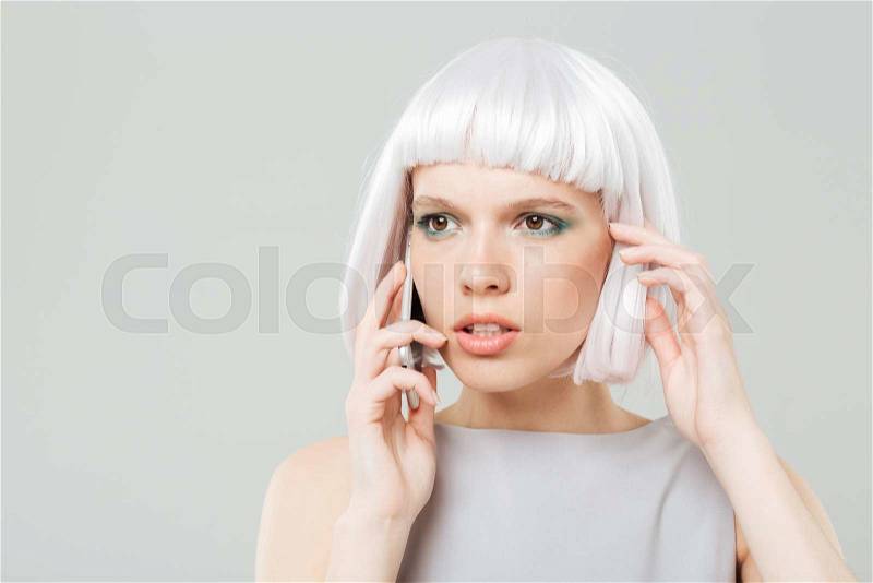 Thoughtful worried young woman in blonde wig talking on mobile phone, stock photo