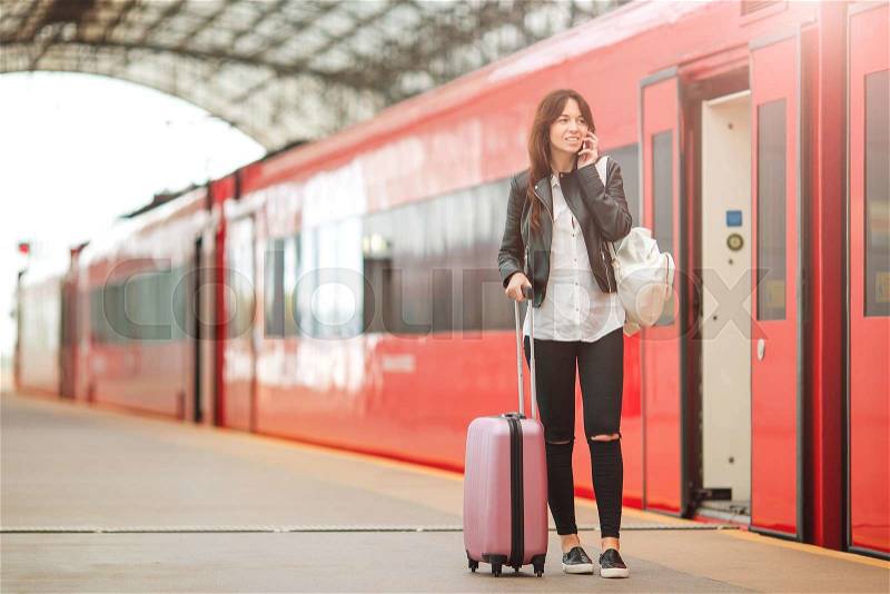 Young woman with luggage talking by cellphone at a train station. Caucasiam tourist waiting her express train while her traveling, stock photo