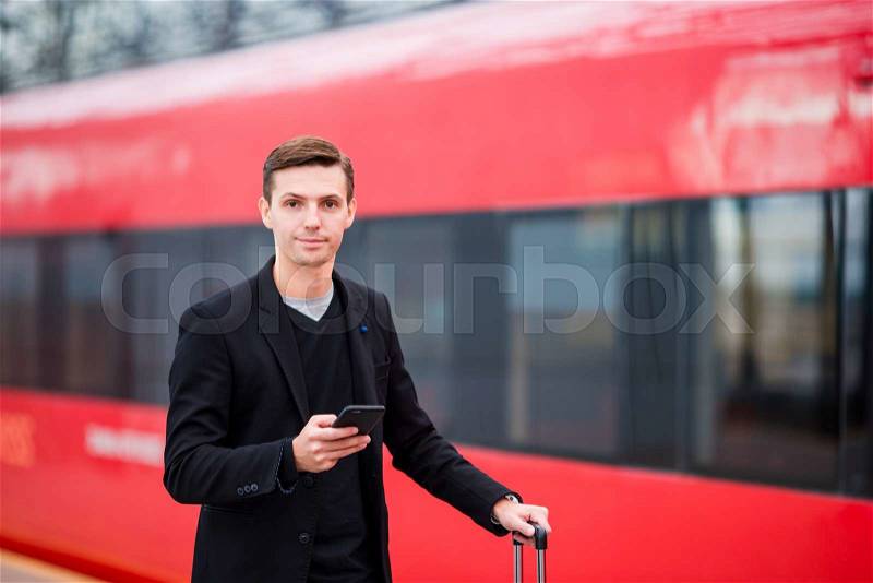 Young caucasian man with smarphone and luggage at station traveling by train, stock photo