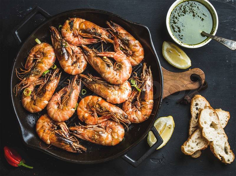 Roasted tiger prawns in iron grilling pan with fresh leek, lemon, bread and pesto sauce over black background, top view, stock photo