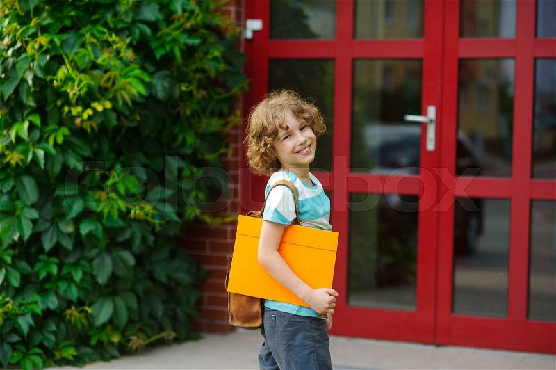 Cheerful pupil coming to the school door. Behind shoulders at the pupil a satchel, in hands the bright folder. The schoolboy with a smile looks in a camera. Back to school, stock photo
