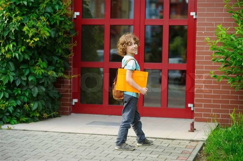 Cheerful first grader coming to the school door. Behind shoulders at the pupil a satchel, in hands the bright folder. The schoolboy with a smile looks in a camera. Back to school, stock photo