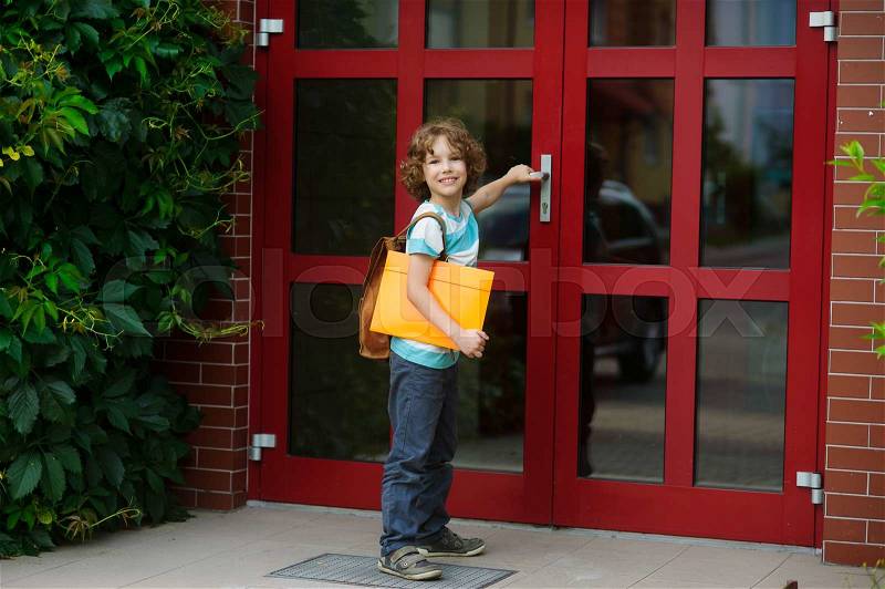 The pupil of elementary school opens a school door. Behind shoulders at the school student a school backpack, in hands the bright folder. The boy with a smile looks in the camera. Back to school, stock photo