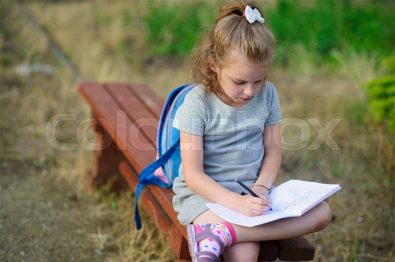 Little school miss sits having crossed legs on a park bench and something diligently writes in the notebook. Girlie has a thoughtful look. Behind the back the school backpack lies. Back to school, stock photo