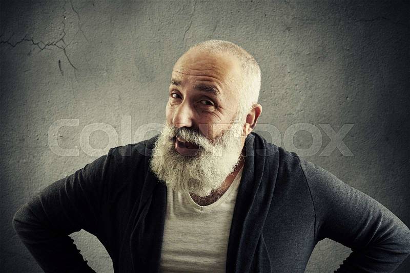 Smiling aged man with white beard in stylish casual clothes is posing at the camera with hands on hips against grey concrete wall, stock photo