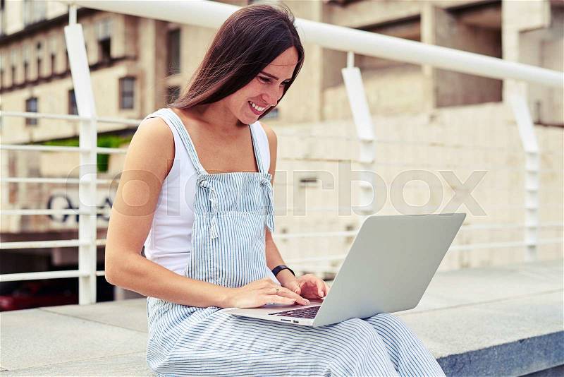Close-up of young attractive lady in overalls working on laptop against city background, stock photo