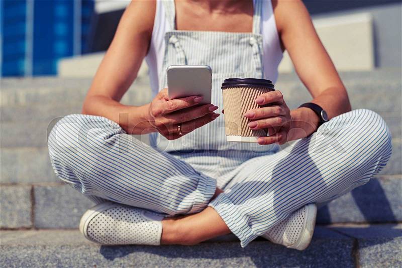 Close-up of slim woman sitting with crossed legs and holding a cup of coffee in one hand and smart phone in another, stock photo