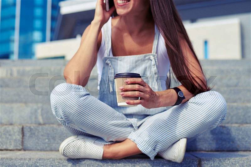 Close-up of young stylish woman sitting with crossed legs and talking on the phone with a cup of coffee in her hands, stock photo