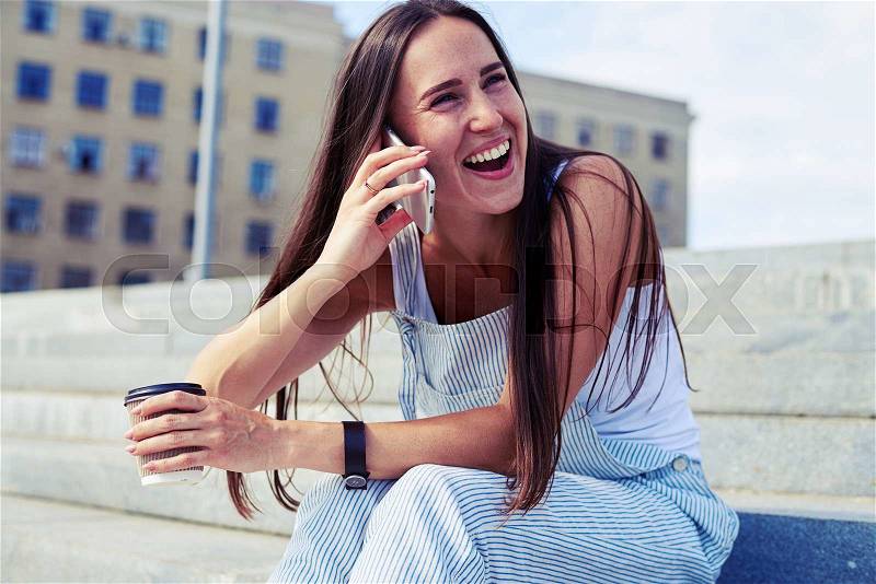 Sexy stylish female is laughing on a joke during her phone talk while having a coffee break in the street on a sunny day, stock photo