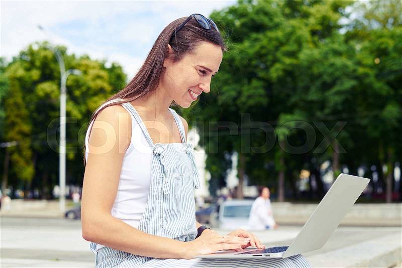 Pretty lady in casual overalls is smiling and using her notebook computer while sitting in the recreational area of the busy city, stock photo