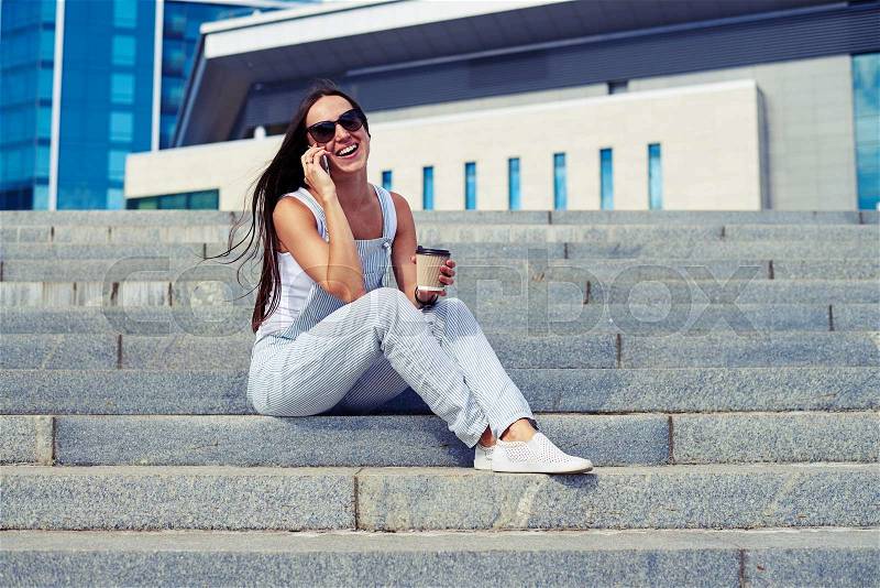 Young attractive smiling lady is sitting on the city stairs with a cup of “coffee to go” and talking on the phone, stock photo