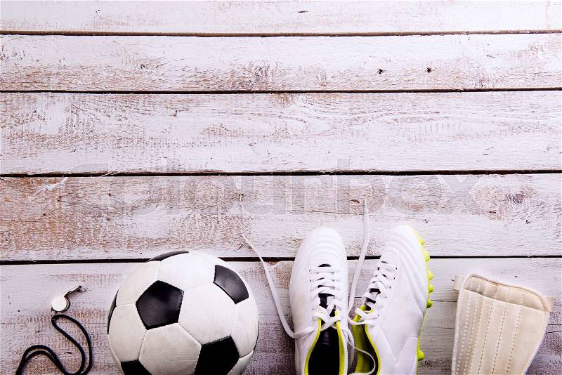 Soccer ball, cleats, protectors and whistle against wooden floor, studio shot on white background. Flat lay, copy space, stock photo