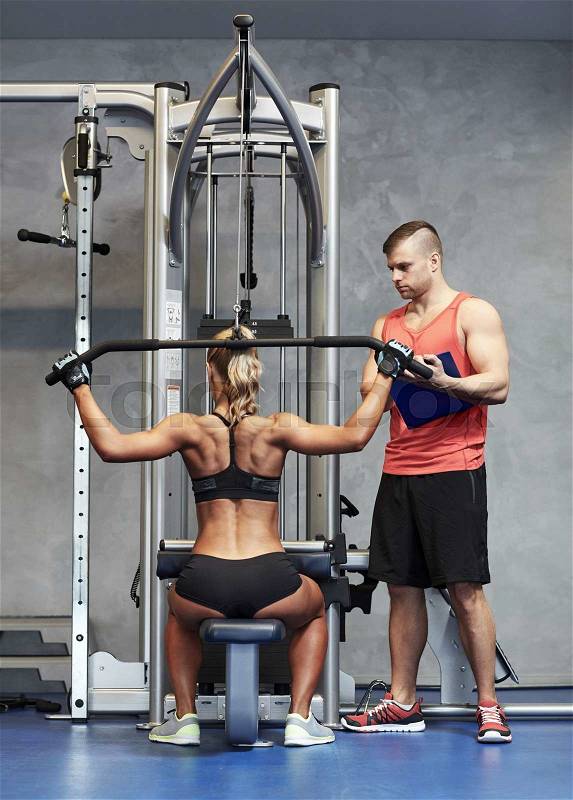 Sport, fitness, teamwork and people concept - young woman and personal trainer flexing muscles on gym machine, stock photo