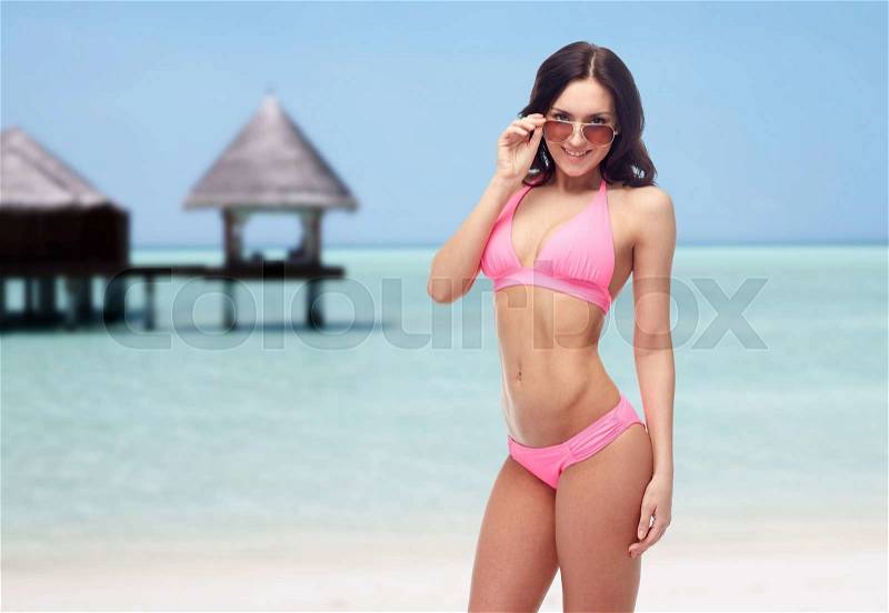 People, fashion, swimwear, summer and travel concept - happy young woman in sunglasses and pink swimsuit looking at you over maldives beach with bungalow background, stock photo