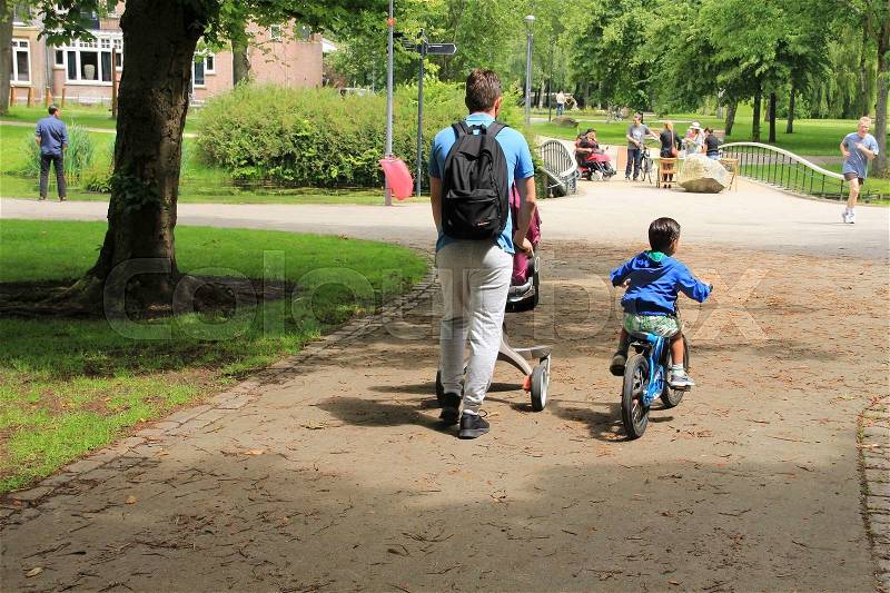The father is walking behind the pram and the little son is biking in the wonderful park in the city Rotterdam in the summer, stock photo