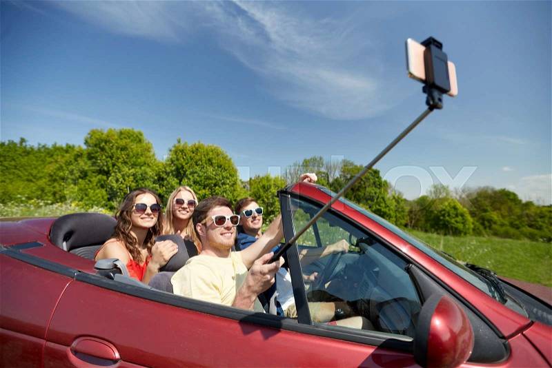 Leisure, road trip, travel and people concept - happy friends driving in cabriolet taking picture by smartphone selfie stick at country, stock photo