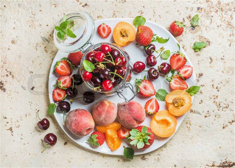 Healthy summer fruit variety. Sweet cherries, strawberries, peaches, apricots and mint leaves on white ceramic serving plate over light concrete background. Top view, selective focus, stock photo