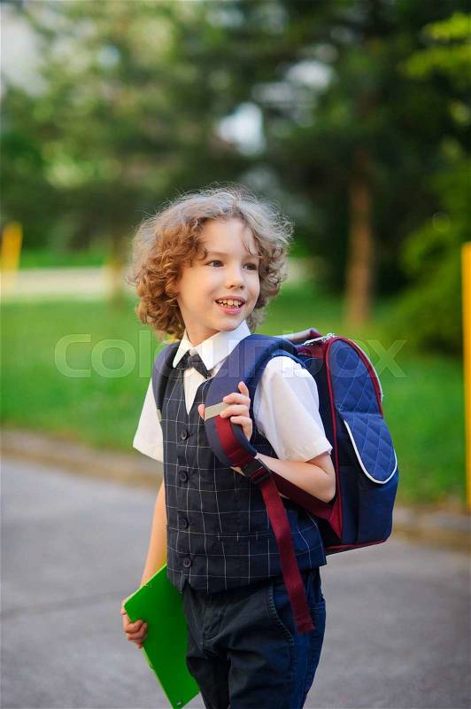 Little schoolboy standing in the school yard. Elementary school student is smartly dressed. Behind the boy\'s school backpack. The first grader looks away and smiles. Back to school, stock photo