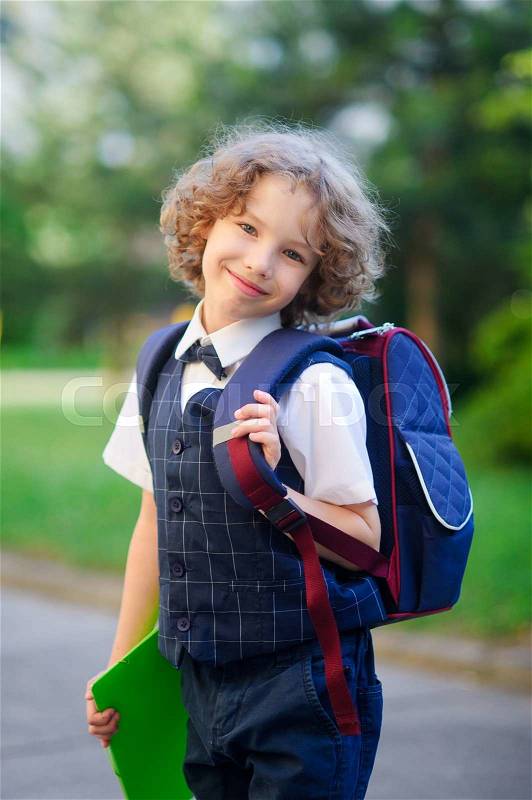 Cute little first grader is in the school yard. The primary school students smartly dressed. Behind the boy\'s school backpack in his hands - bright folder. The schoolboy looks at camera and smiling. Back to school, stock photo