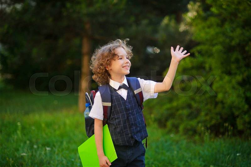 Curly blonde first-grader goes to school. The primary school students smartly dressed. Behind the boy\'s school backpack. The schoolboy looks to the side and someone waving his hand. Back to school, stock photo