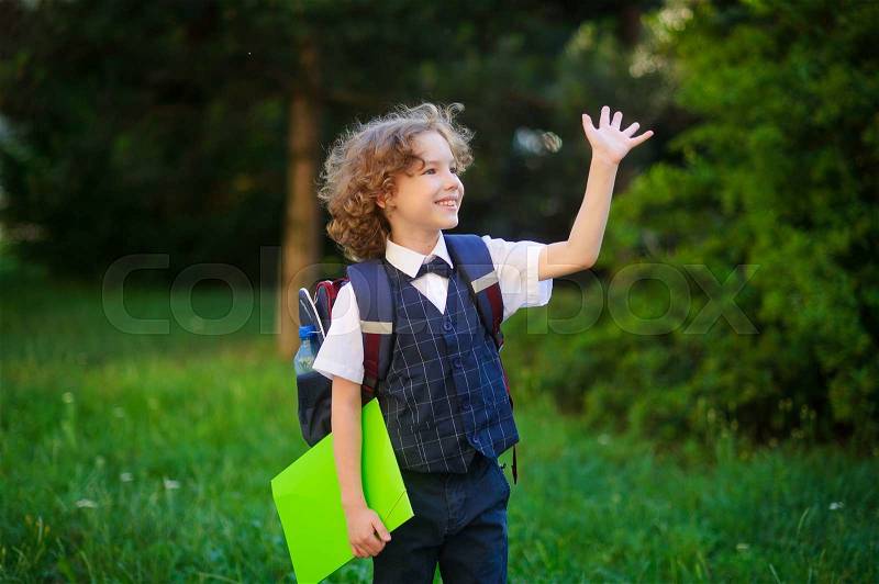 Blonde first-grader goes to school. The primary school students smartly dressed. Behind the boy\'s school backpack in his hands - bright folder. Schoolboy looks to the side and someone waving his hand, stock photo