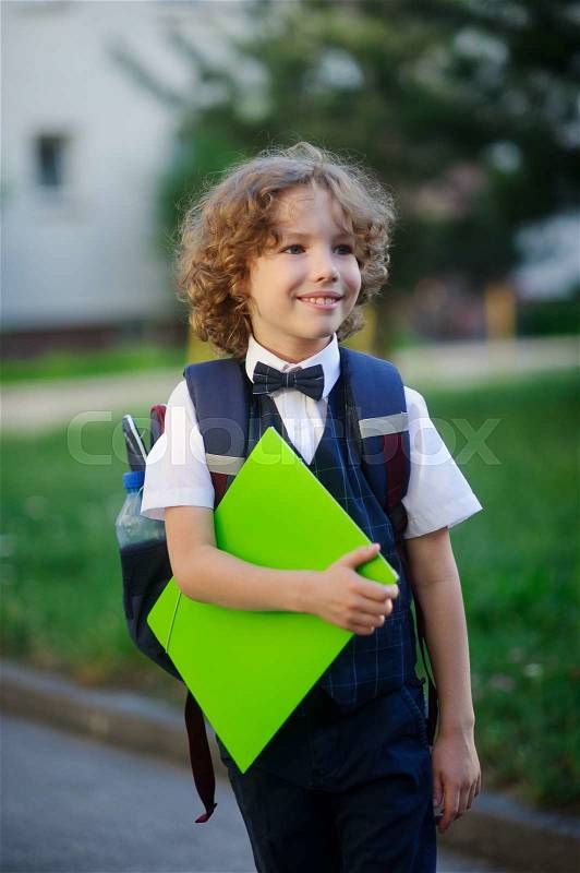 Elegant fair-haired first-grader in the school yard. The student looks at the camera with a sweet smile. In hand the boy has a bright green folder. Behind him a school bag. Back to school, stock photo