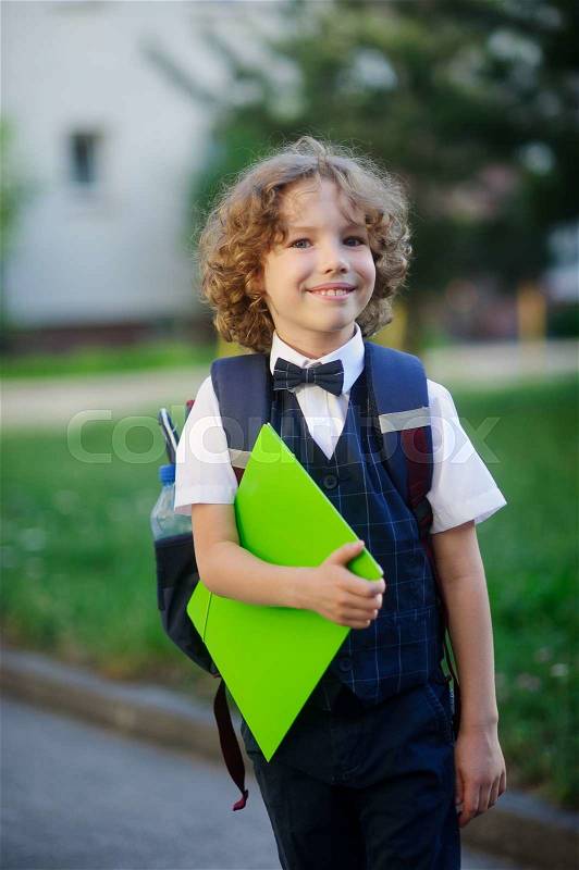 Elegant fair-haired first-grader in the school yard. The student looks at the camera with a sweet smile. In hand the boy has a bright green folder. Behind him a school bag. Back to school, stock photo