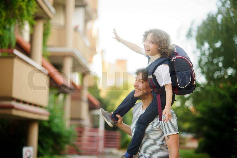 Little pupil sitting on the shoulders of his father. He placed his hands to the sides as if flying. Dad and son a great mood. They play on the way to school. Behind the schoolboy\'s backpack, stock photo