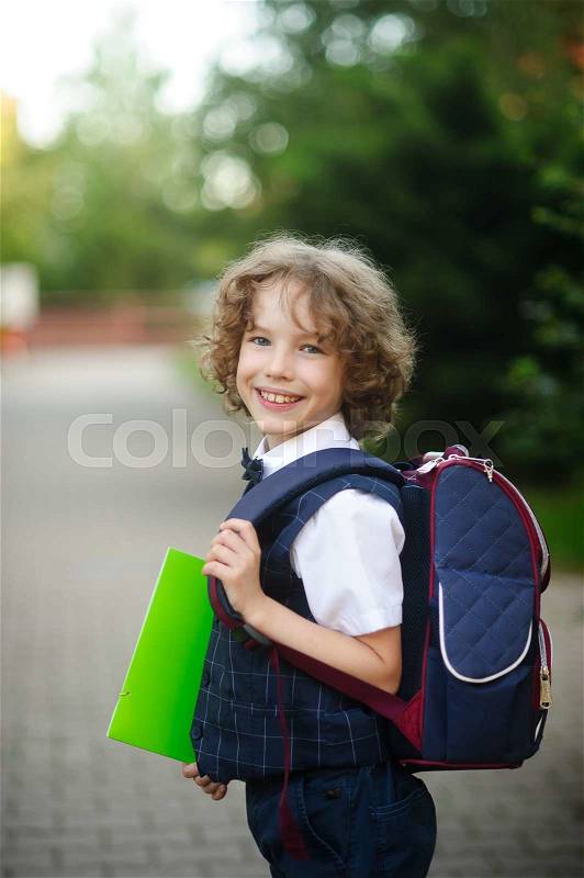 Little curly grader stands in the school yard. The boy\'s sweet face. Behind backpack in hands - bright green folder. Schoolboy looks into the camera and smiling, stock photo