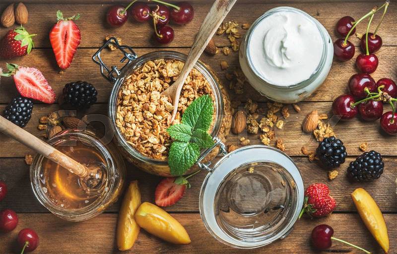 Healthy breakfast ingredients. Oat granola in open glass jar, fruit, yogurt, berries and mint over wooden background, top view, stock photo