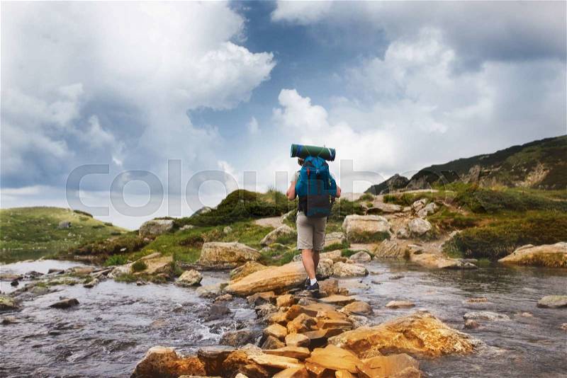Hiker man with backpack crossing a river on stones in bulgarian mountains. Hiking and leisure theme. Image with sunlight effect, stock photo