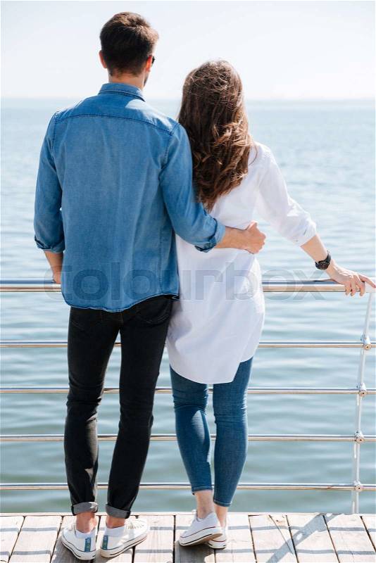 Full length of a young couple standing backwards hugging and looking ahead at the seaside, stock photo