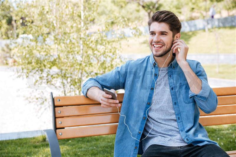 Portrait of a happy man sitting on the bench with laptop computer and talking on the phone outdoors, stock photo
