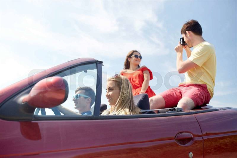 Leisure, road trip, travel, summer holidays and people concept - happy friends driving in cabriolet car and taking picture by film camera, stock photo