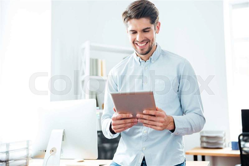 Cheerful young businessman standing and using tablet in office, stock photo