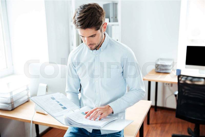 Concentrated young man working and holding folder with documents in office, stock photo