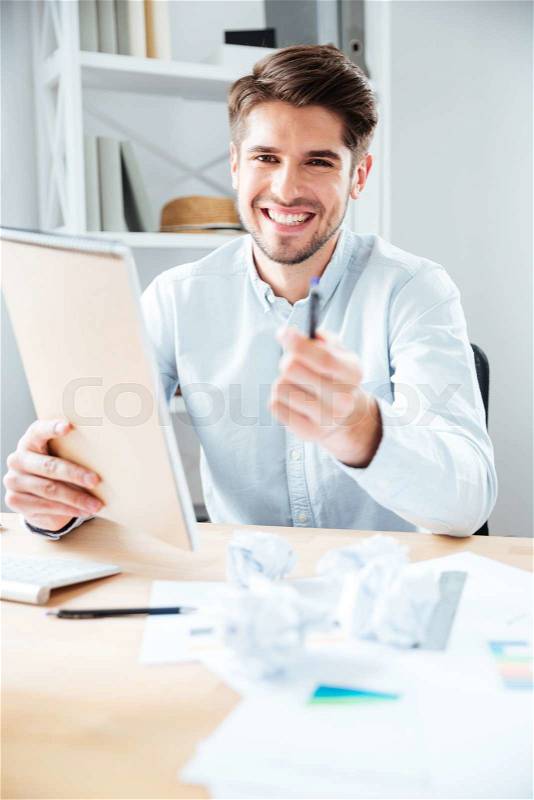 Cheerful young businessman giving notepad and pen to you in office, stock photo