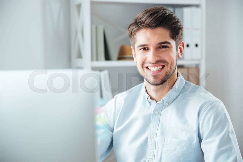 Happy young businessman using laptop computer in office and looking at camera, stock photo