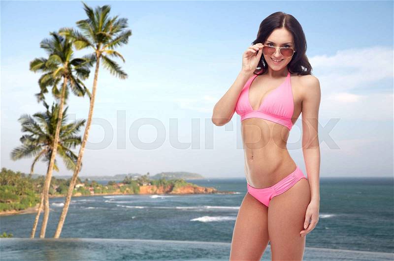 People, fashion, swimwear, summer and travel concept - happy young woman in sunglasses and pink swimsuit looking at you over Sri Lanka beach with palms swimming pool background, stock photo