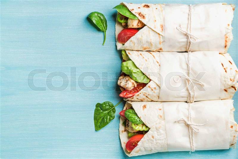 Healthy lunch snack. Three tortilla wraps with grilled chicken fillet and fresh vegetables over turquoise blue painted wooden background. Top view, copy space, horizontal composition, stock photo