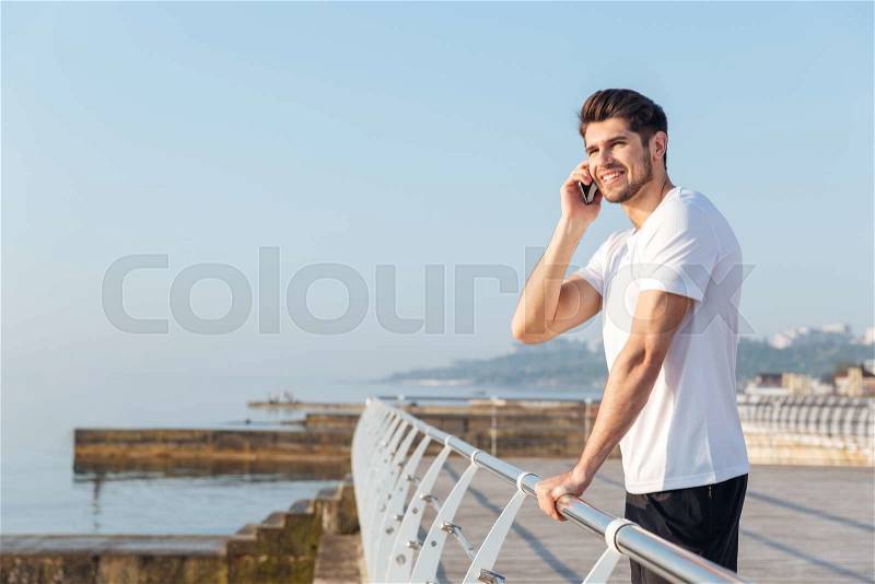Cheerful young sportsman standing and talking on mobile phone outdoors, stock photo