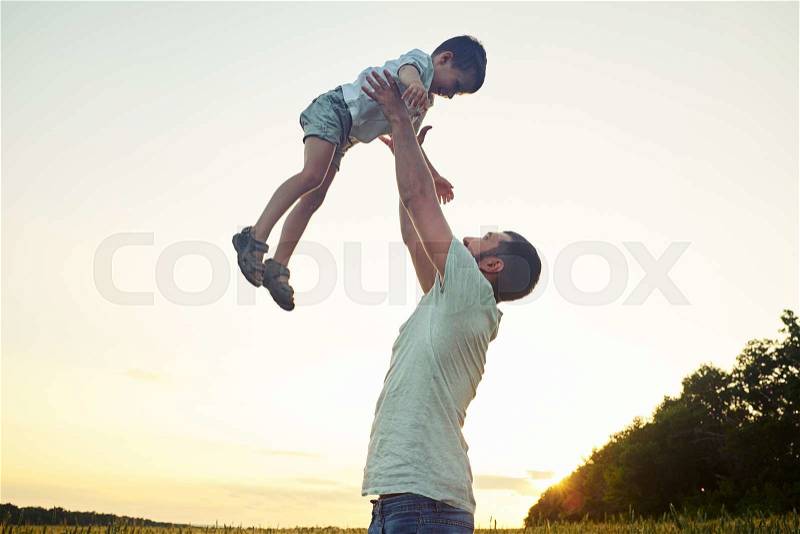 Happy father is playing with his small son lifting him in the air on the background of beautiful sunset sky, stock photo