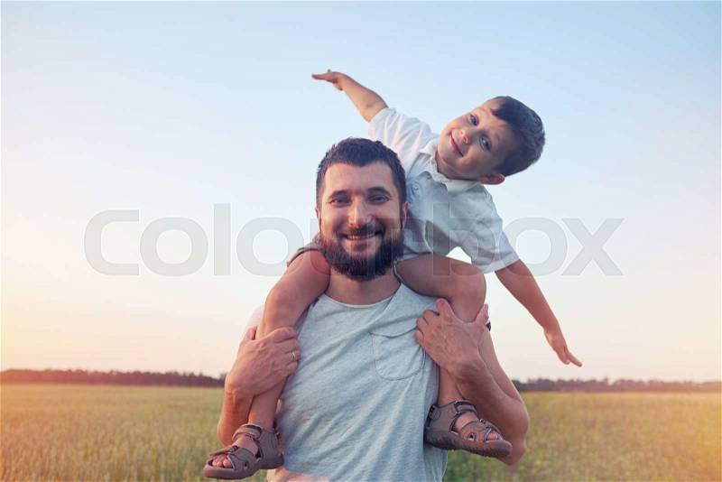 Small boy is sitting on his father\'s shoulders and pretending a plane during walk in the field, stock photo