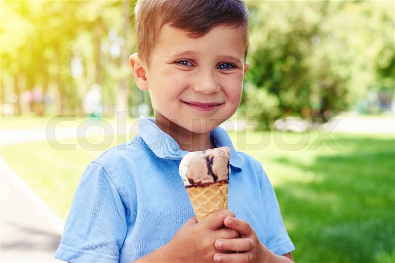Small boy with beautiful blue eyes is smiling and holding an ice-cream in the park on a warm sunny summer day, stock photo