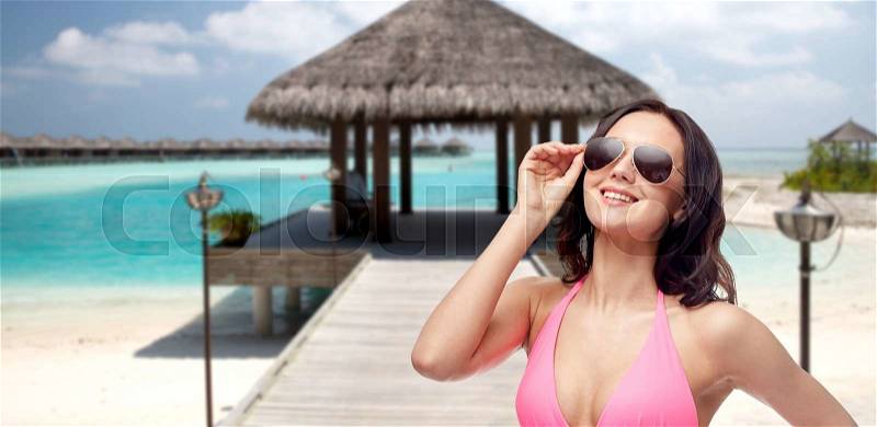 People, fashion, swimwear, summer and travel concept - happy young woman in sunglasses and pink bikini swimsuit over maldives beach with bungalow background, stock photo