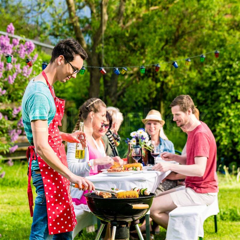 Man grilling meat on garden barbecue party, in the background friends eating and drinking, stock photo