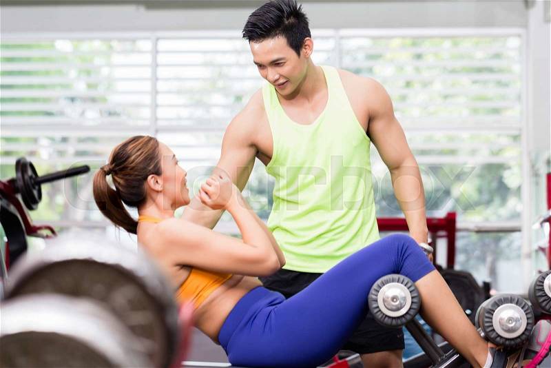 Sportive male instructor advising Asian woman doing sit-ups on bench in gym , stock photo