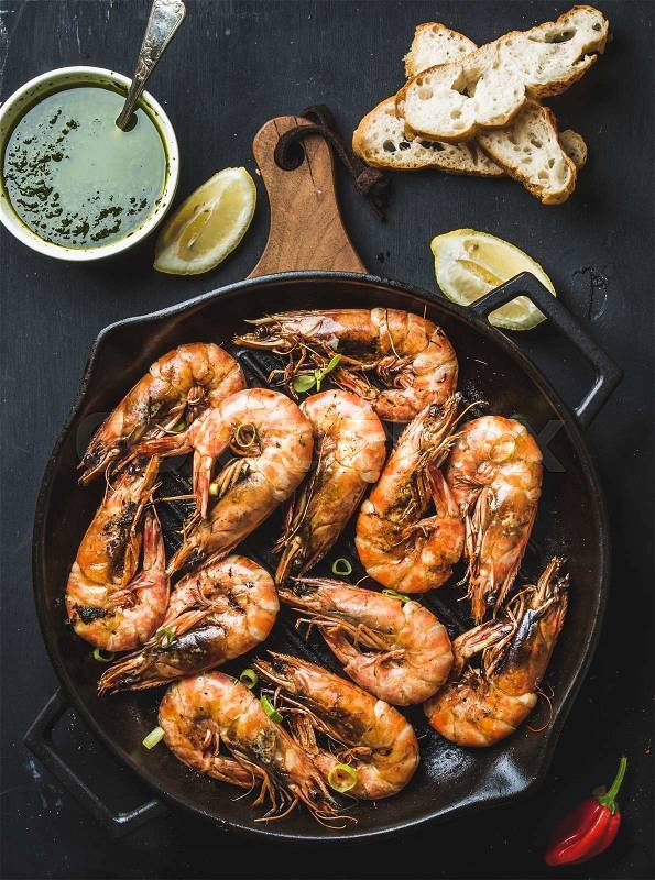 Roasted tiger prawns in iron grilling pan on wooden board with fresh leek, lemon slices, bread, pepper and pesto sauce over black background, top view, vertical composition, stock photo