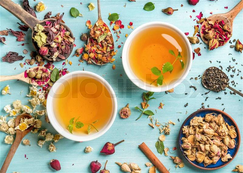 Two cups of healthy herbal tea with mint, cinnamon ,dried rose and camomile flowers in spoons over blue background, top view, stock photo